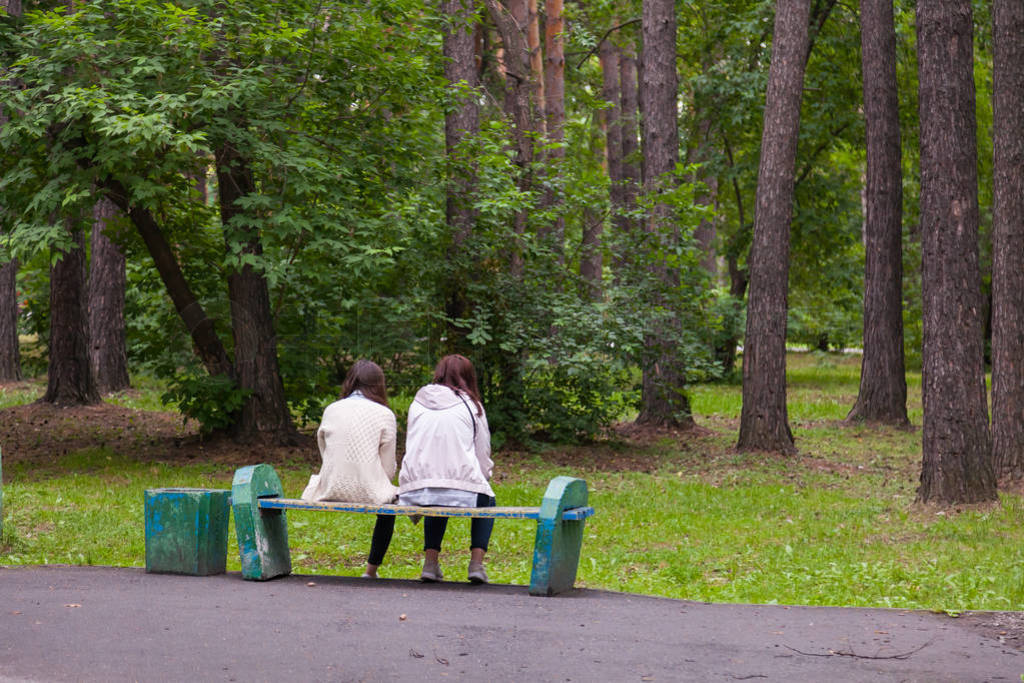 Two girls girlfriends sit on an old wooden bench in a city park