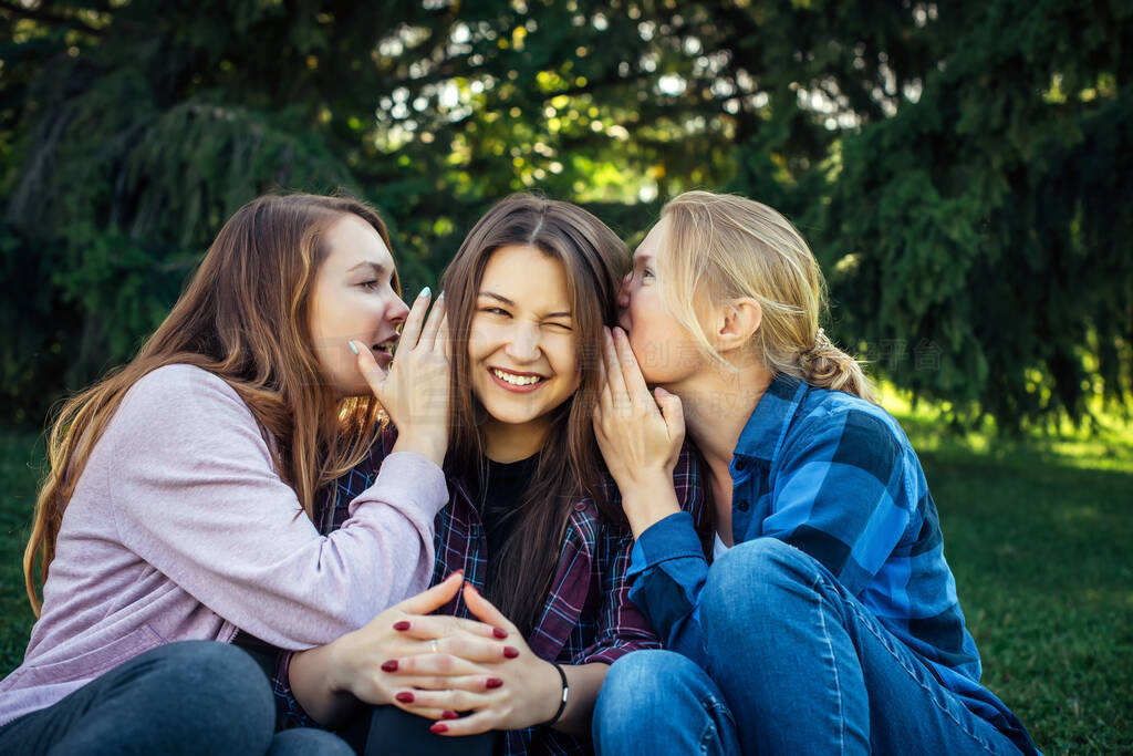 Three young attractive woman sharing secrets sitting on green gr