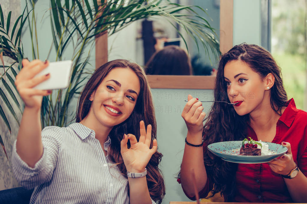 Two girl eating cakes and taking selfie with smartphone in cafe