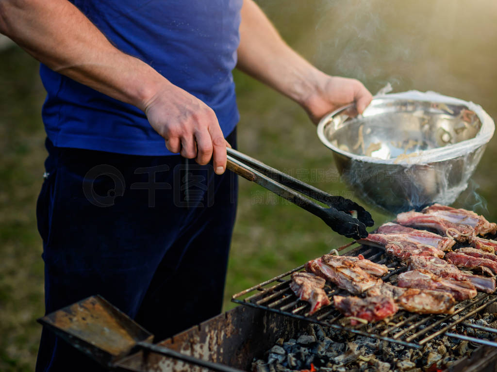 Man's hand with metal tongs turns the lamb pieces on the gril