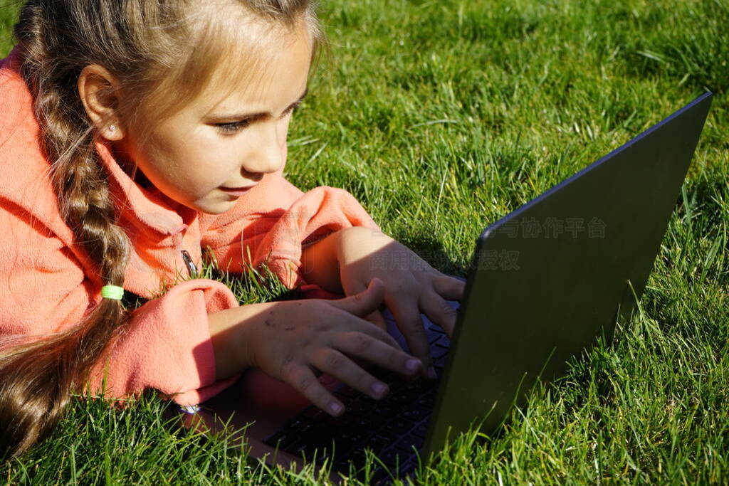 A girl of seven years old works on a laptop on the green grass.