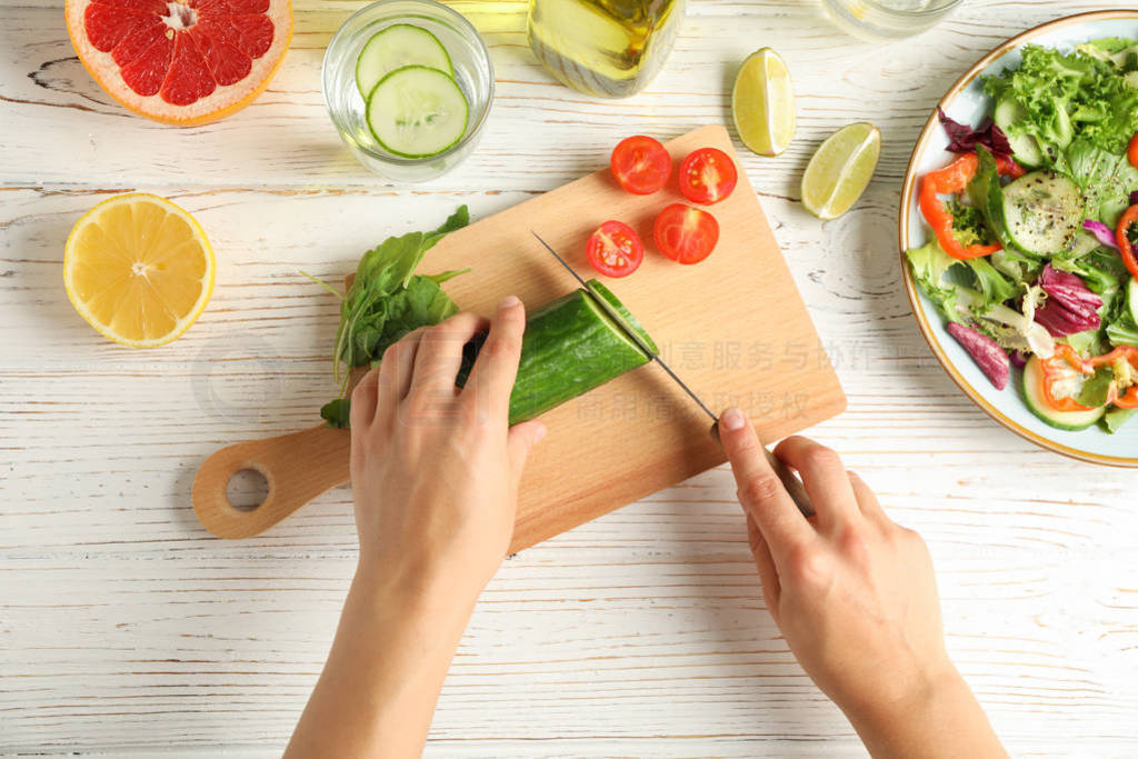 Young woman slices cucumber for salad on wooden background, top