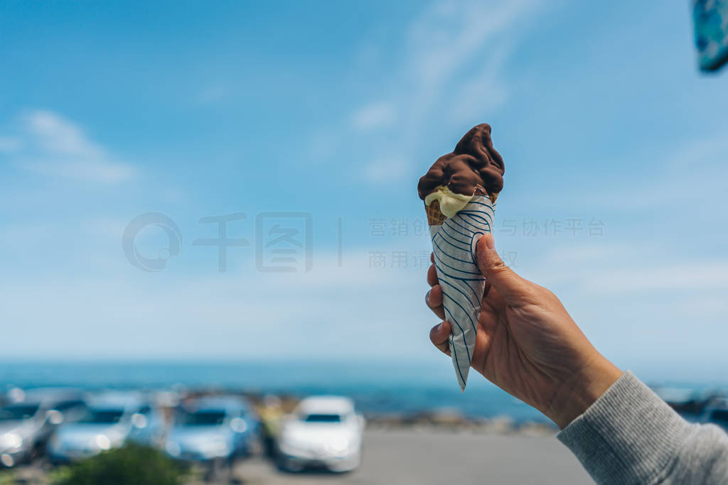 Man's hand holds ice cream in cone against blue sky.