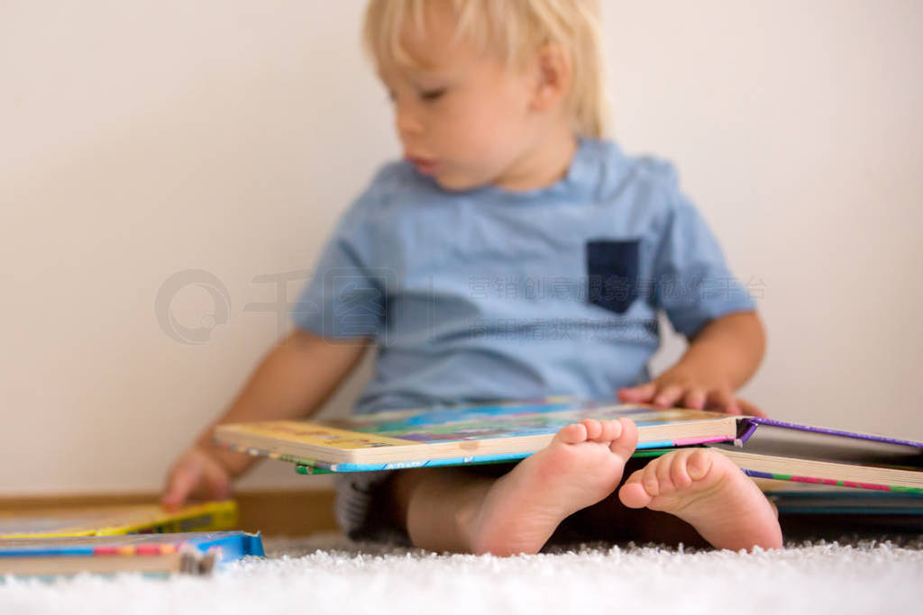 Sweet toddler boy, reading book at home, sitting on the ground,