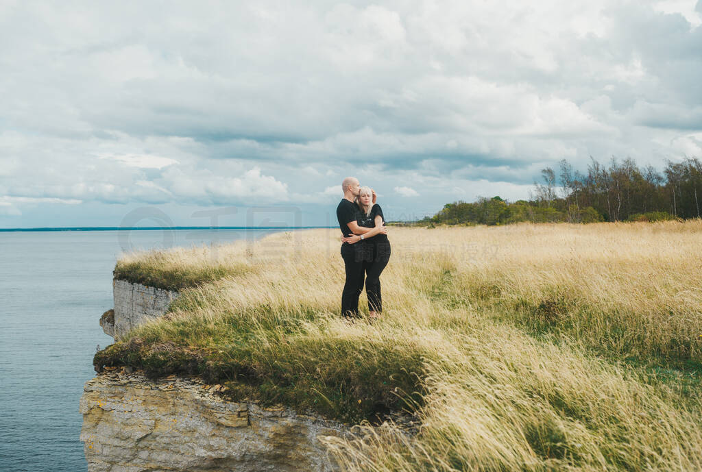 A couple embrace on grass on the edge of the rocky coast of the