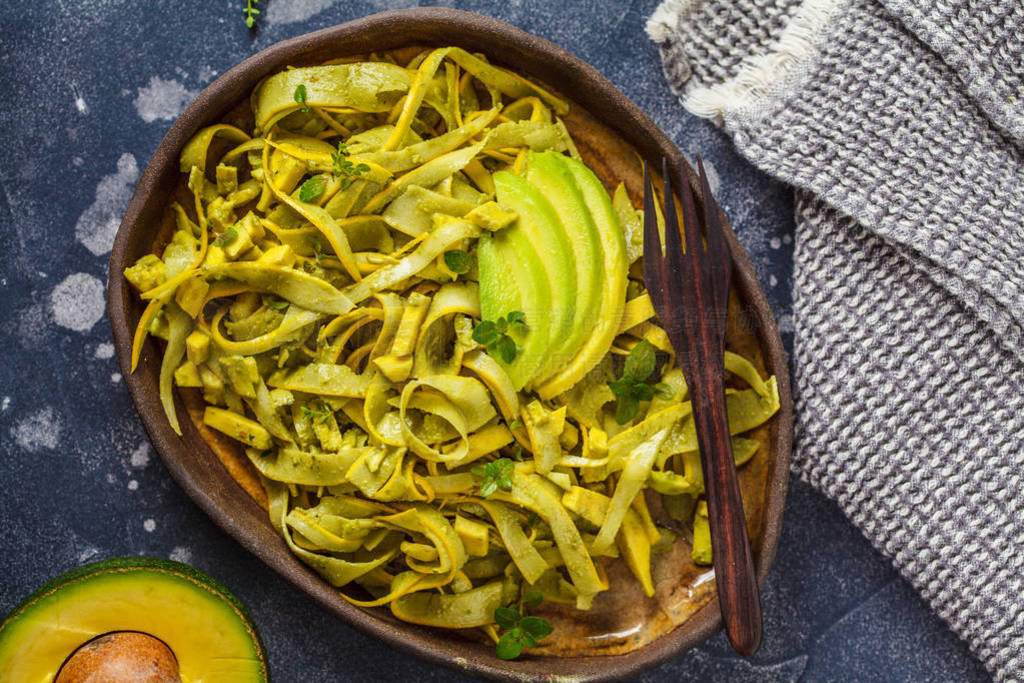 Zucchini pasta with pesto and avocado in dark dish.