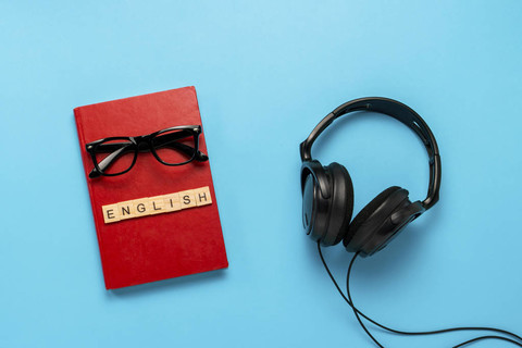 带有红色封面的书，上面有文字英语眼镜和蓝色背景上的黑色耳机。 有声读物的概念，自主教育和学习英语。 平躺顶部视图。