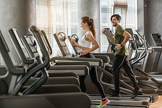 健康的年轻男女在健身房的现代电动跑步机上并肩奔跑时微笑的侧<i>视</i>图