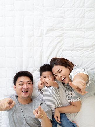 可爱的小男孩和他的亚洲父母躺在家里的白<i>床</i>上微笑着用手指指着某物的顶视图 幸福家庭的概念