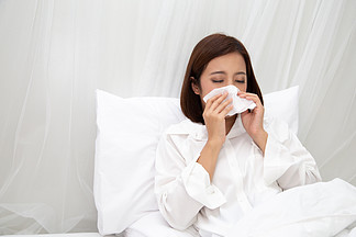 年轻的亚洲女性在卧室的白床上用纸巾打<i>喷</i>嚏 流感和过敏概念