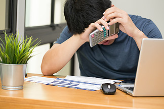 亚洲男人在用计算器计算信用卡债务并在家里用笔记本电脑工作时强调 <i>悲</i><i>伤</i>和戏剧性 债务压力和心理健康概念