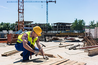 一名身穿安全背心和黄色安全帽的年轻工人在当代建筑工地工作时将钉子钉入木头的侧<i>视</i>图