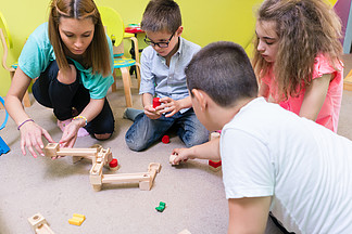 一位<i>敬</i><i>业</i>的幼儿园老师在有监督的免费游戏时间帮助孩子们建造木制火车线路的低角度视图