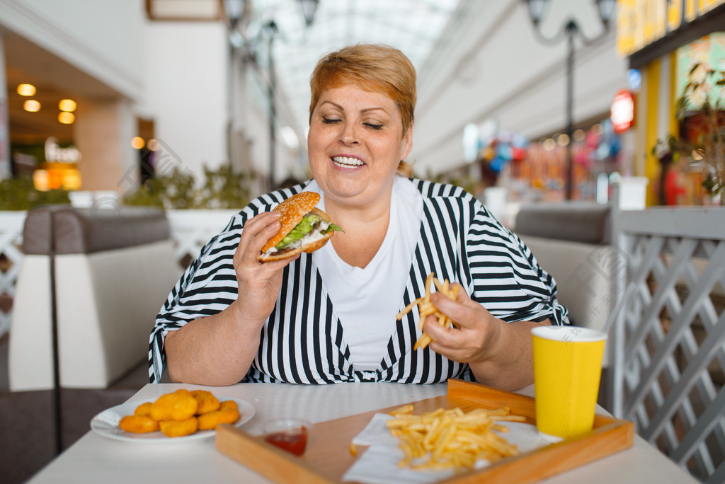 吃高卡路里食物的肥胖妇女在快餐餐馆超重的女性在桌上用垃圾晚餐