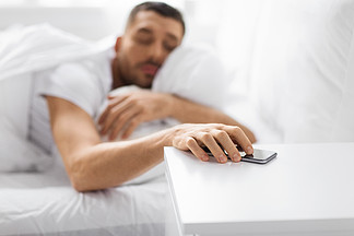 技术和人概念-接近早晨在家<i>到</i>达为在床头柜上的智能手机的困年轻人从床困了年轻男子伸手躺在床上的智能手机
