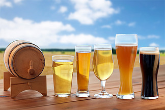啤酒厂，饮料和酒精的概念— —不同类型的啤酒在眼镜和桶在麦田和蓝蓝的天空背景上的桌子上不同类型的啤酒在桌上的眼镜