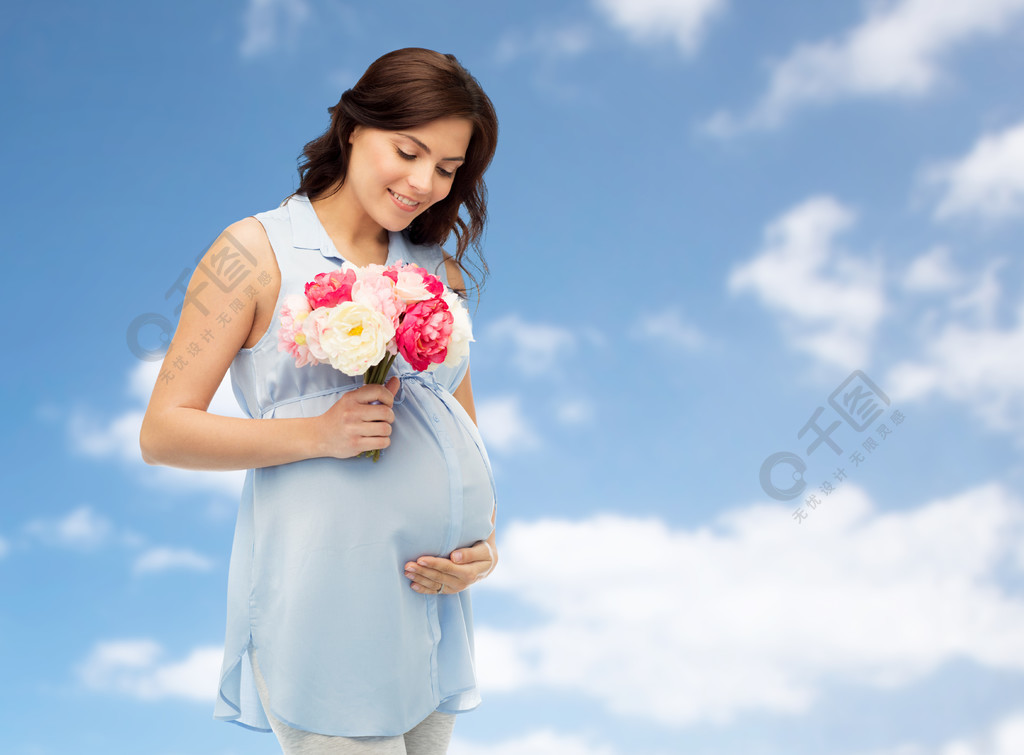 幸福的孕妇,与花抚摸她的大肚子在蓝蓝的天空和云彩背景