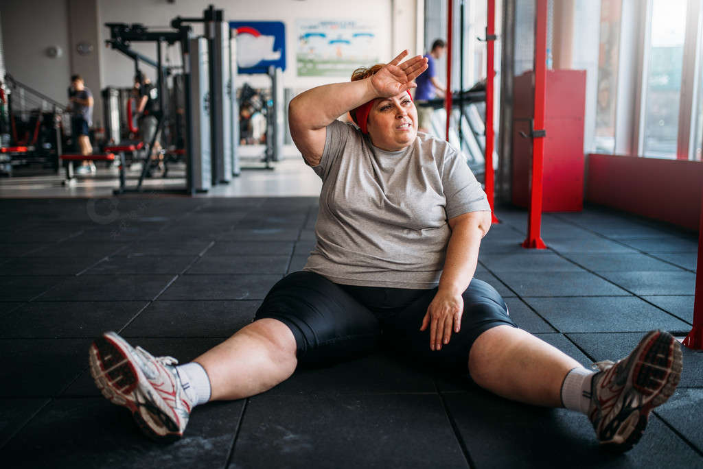 超重女人坐在健身房的地板上卡路里燃烧,运动俱乐部中的肥胖女性,胖子