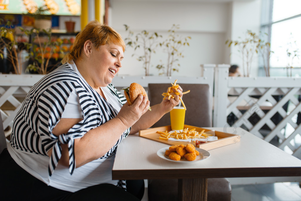 吃高卡路里食物的肥胖妇女在快餐餐馆超重的女性人在桌上的垃圾晚餐