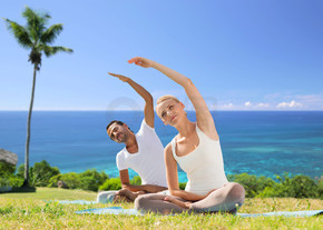 健身，体育和人们的观念-夫妇在户外做瑜伽运动与棕榈树和海洋的异国自然背景幸福的夫妻，在户外做瑜伽练习