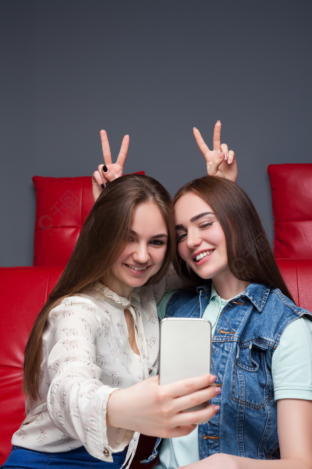 两个有趣的女朋友坐在红色的皮革沙发上,使手机相机拍照女性的友谊
