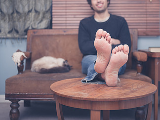 一个赤脚的年轻人在家里的咖啡桌上休息他的双腿，他旁边的沙发上有只猫