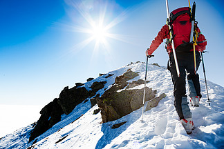登山者沿着陡峭的白雪皑皑的山脊与背包里的滑雪板走在背景中戏剧性的天空与闪亮的灿烂的阳光概念：冒险，成就，勇气，决心，自我实现，危险活动，极限运动，冬季休闲