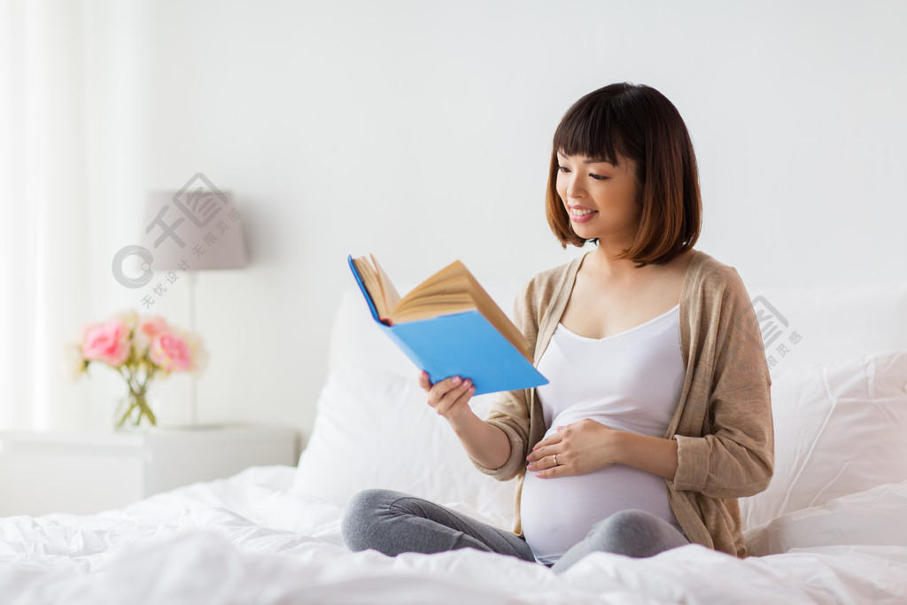 微笑的怀孕的亚裔妇女阅读书在床上在家幸福的怀孕亚洲女人在家里读书
