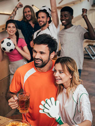 快乐的年轻夫妇和多文化的一群朋友庆祝和手势在观看足球比赛在酒吧