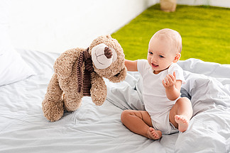 有趣的赤脚孩子在白色的衣服坐在床上，抱着毛绒熊