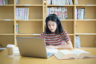 一个女大学生坐在桌旁做大学图书馆的作业。 学生使用笔记本电脑，在网上学习.