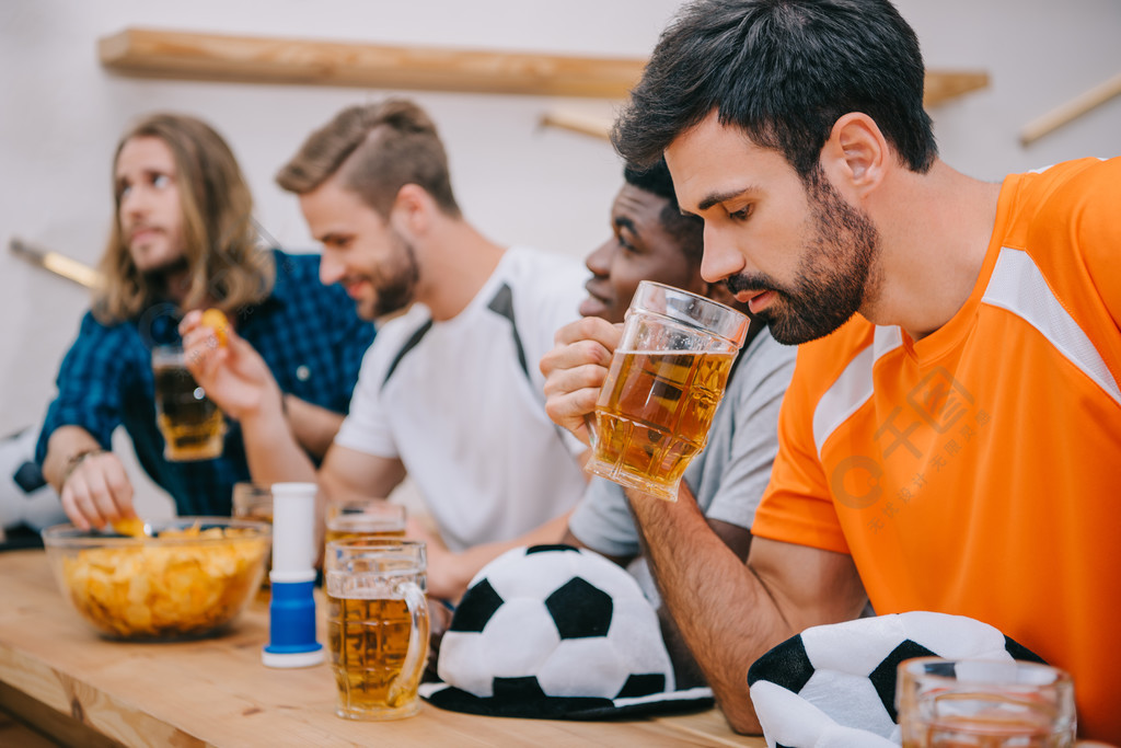 沮丧的年轻人在橙色 t恤的侧面观看啤酒, 而他的朋友坐在酒吧柜台与球帽, 薯条, 风扇喇叭和啤酒在观看足球比赛