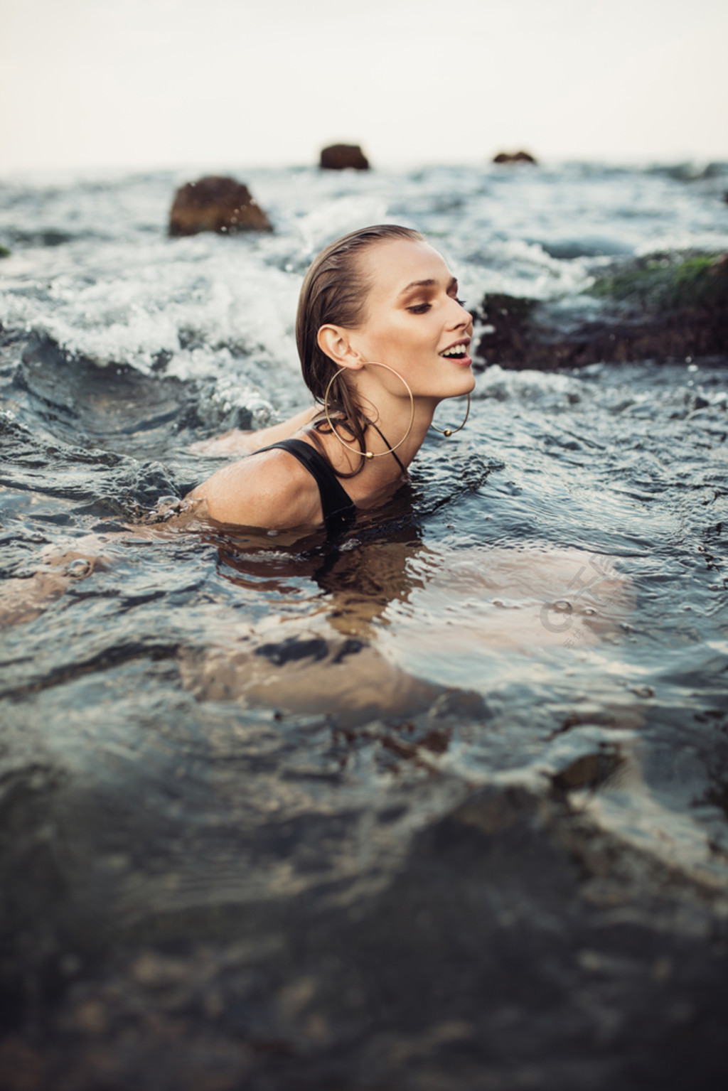 的微笑的女孩与湿头发在黑色泳衣和大现代耳环快乐地独自在海里游泳