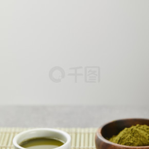 在木碗中选择性聚焦绿色火柴粉，靠近白杯，竹桌垫上茶