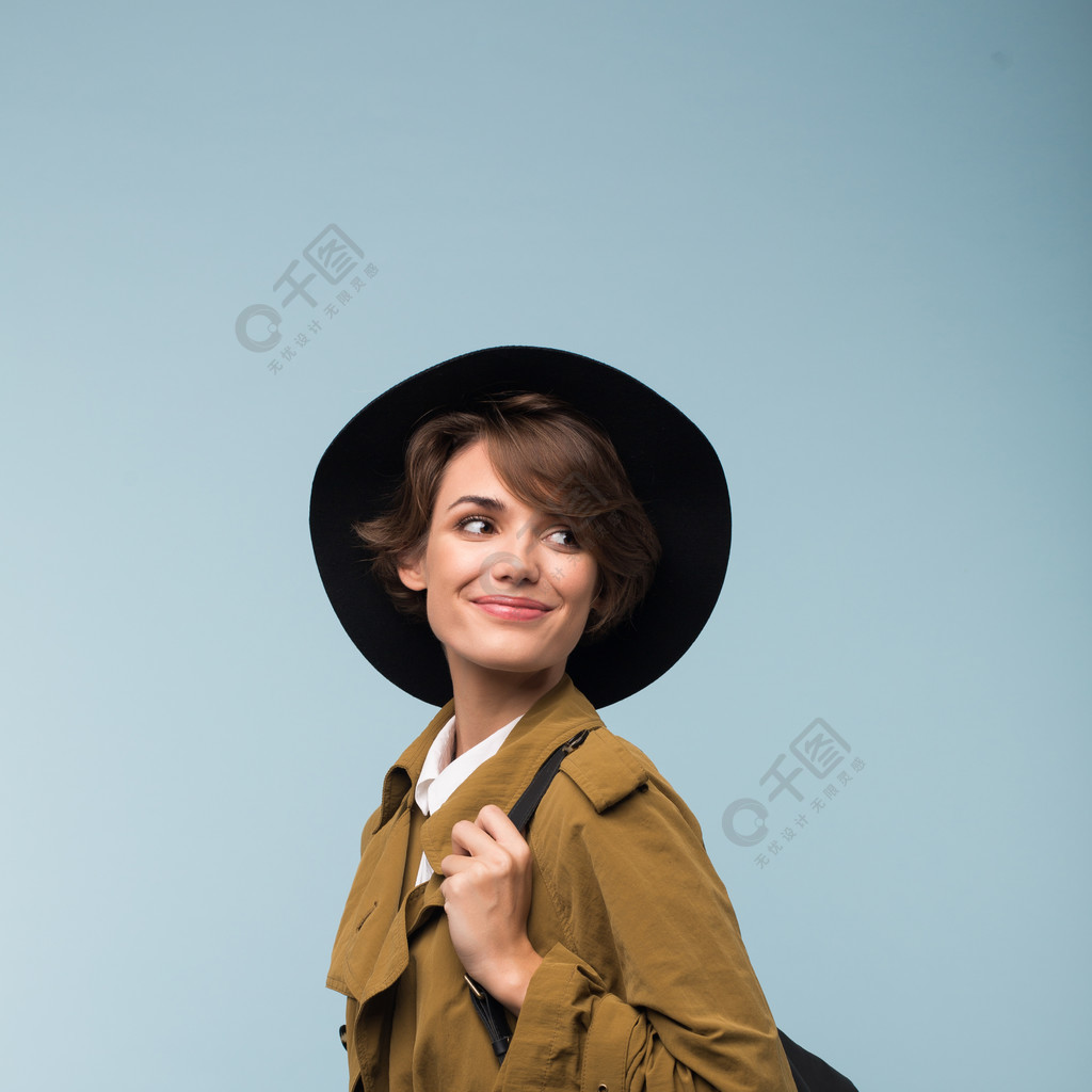 美丽的微笑的女孩与黑暗的短发在风地衣和帽子与背包梦幻般地看在一边在蓝色背景